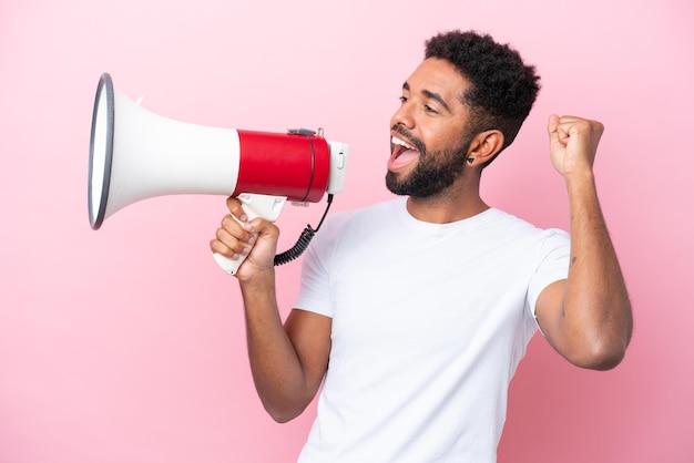 Young Brazilian man isolated on pink background shouting through a megaphone to announce something in lateral position