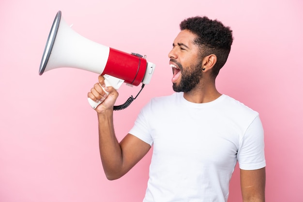 Young Brazilian man isolated on pink background shouting through a megaphone to announce something in lateral position