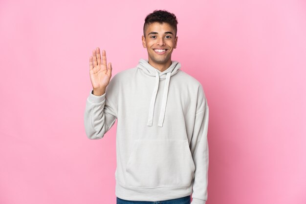 Young Brazilian man isolated on pink wall saluting with hand with happy expression