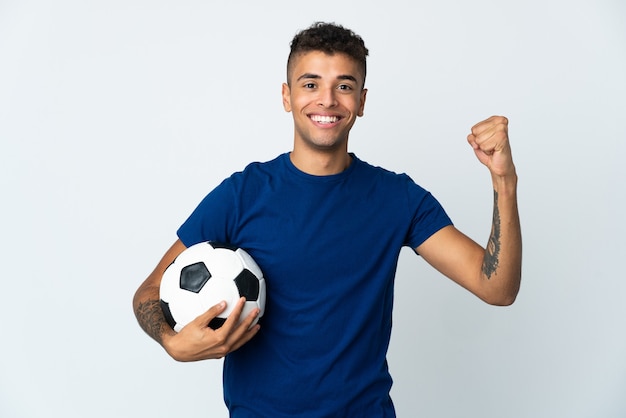 Young Brazilian man over isolated wall with soccer ball celebrating a victory