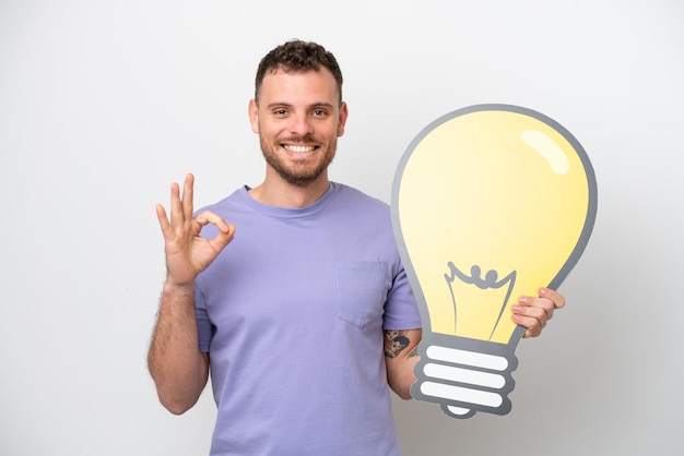 Young Brazilian man isolated on white background holding a bulb icon and doing OK sign