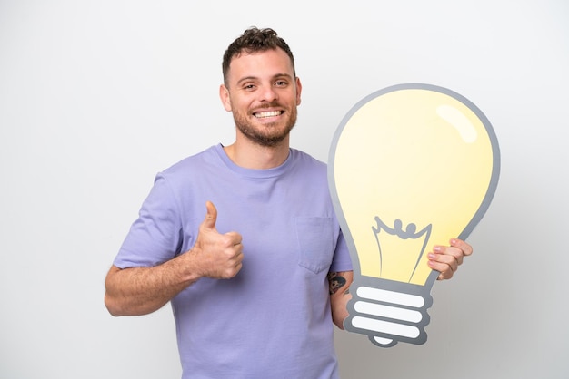 Young Brazilian man isolated on white background holding a bulb icon with thumb up