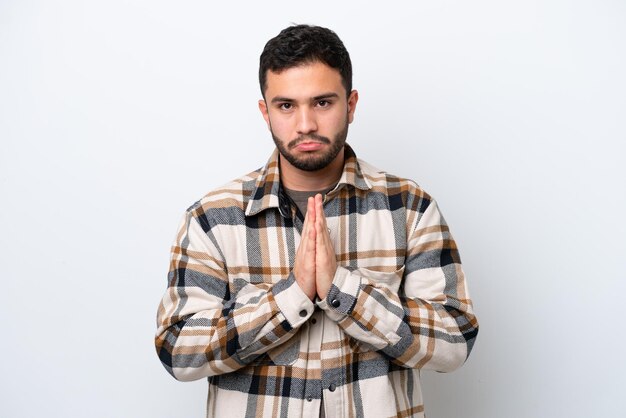 Young Brazilian man isolated on white background keeps palm together Person asks for something