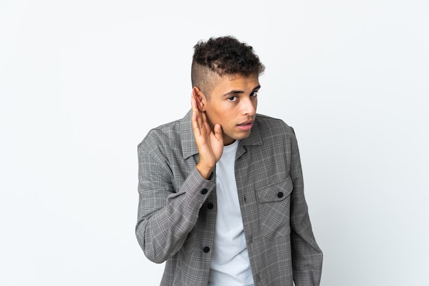 Young brazilian man isolated on white background listening to something by putting hand on the ear