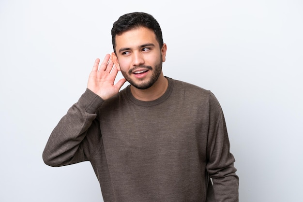 Young Brazilian man isolated on white background listening to something by putting hand on the ear