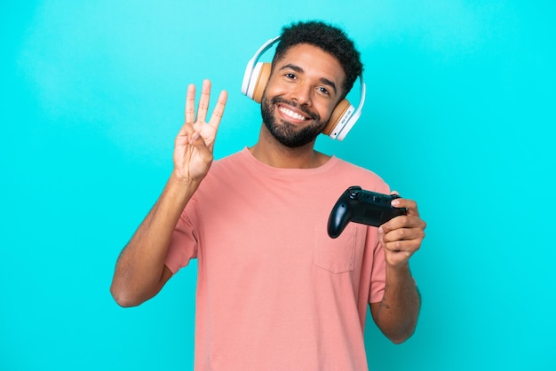 파란색 배경에 격리된 비디오 게임 컨트롤러를 가지고 노는 젊은 브라질 남자가 행복하고 손가락으로 3을 세고 있습니다.