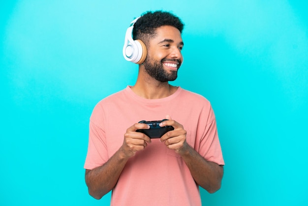 Young brazilian man playing with a video game controller isolated on blue background looking side