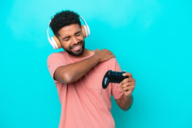 Young brazilian man playing with a video game controller isolated on blue background suffering from pain in shoulder for having made an effort