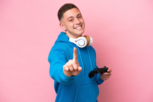 Young brazilian man playing with a video game controller isolated on pink background showing and lifting a finger