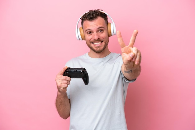 Young Brazilian man playing with video game controller isolated on pink background smiling and showing victory sign