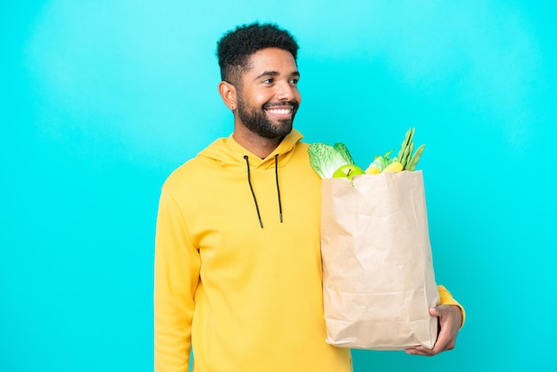 Young brazilian man taking a bag of takeaway food isolated on blue background looking side