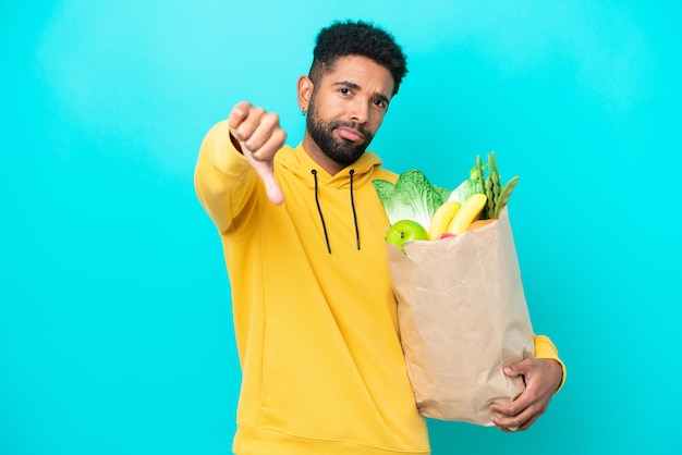 Young Brazilian man taking a bag of takeaway food isolated on blue background showing thumb down with negative expression