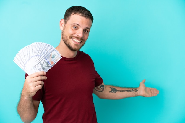 Young Brazilian man taking a lot of money over isolated background extending hands to the side for inviting to come