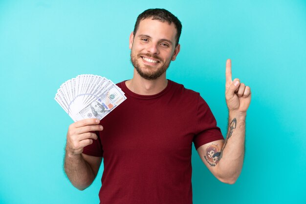 Young Brazilian man taking a lot of money over isolated background showing and lifting a finger in sign of the best