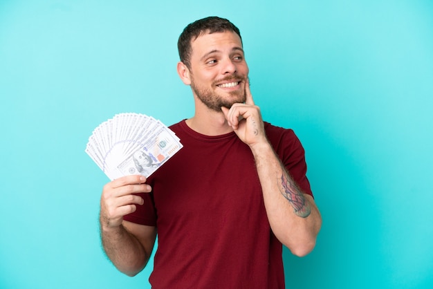 Young Brazilian man taking a lot of money over isolated background thinking an idea while looking up