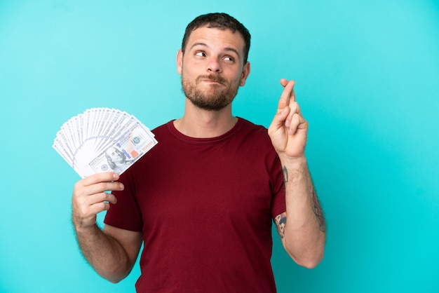 Young Brazilian man taking a lot of money over isolated background with fingers crossing and wishing the best