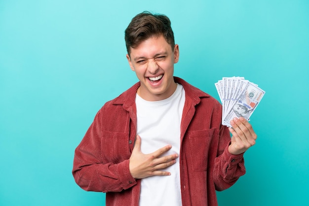 Young brazilian man taking a lot of money isolated on blue background smiling a lot