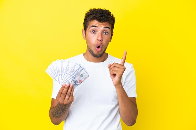 Young brazilian man taking a lot of money isolated on yellow background intending to realizes the solution while lifting a finger up