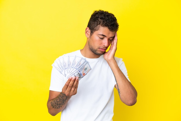 Young brazilian man taking a lot of money isolated on yellow background with headache