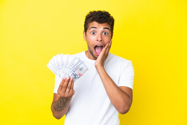 Young brazilian man taking a lot of money isolated on yellow background with surprise and shocked facial expression