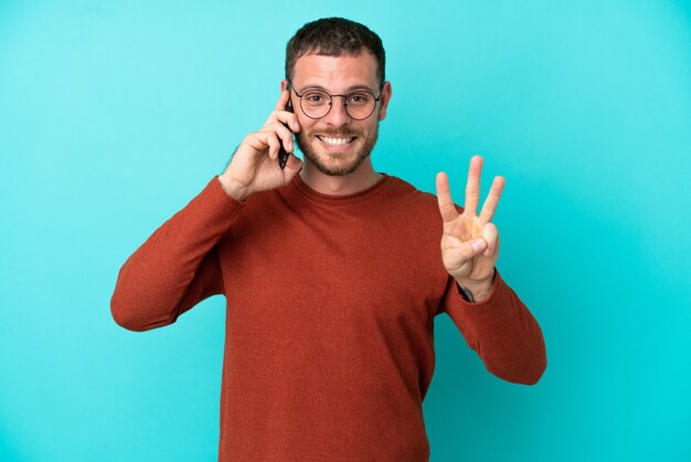 幸せな青い背景に分離された携帯電話を使用して、指で3を数える若いブラジル人男性