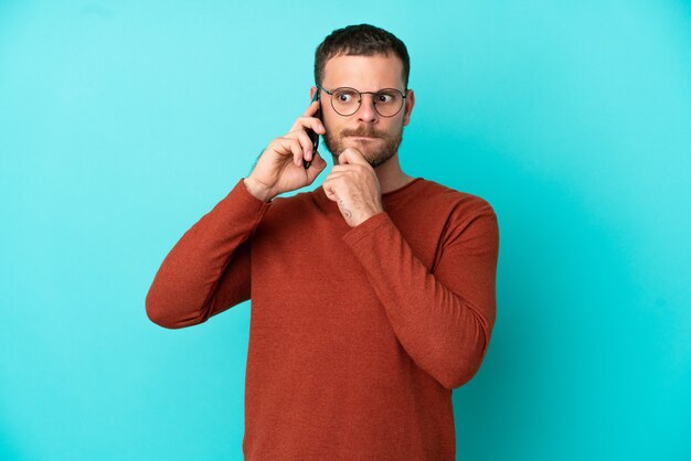 Young Brazilian man using mobile phone isolated on blue background having doubts and thinking