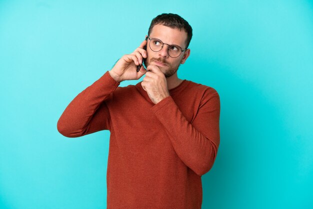 Young Brazilian man using mobile phone isolated on blue background and looking up