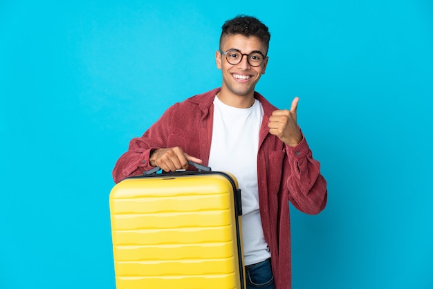 Young Brazilian man over in vacation with travel suitcase and with thumb up