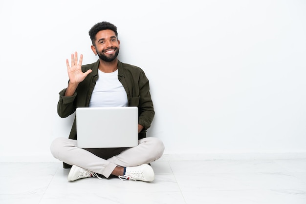 Giovane brasiliano con un computer portatile seduto sul pavimento isolato su bianco contando cinque con le dita