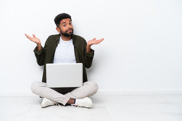 Young Brazilian man with a laptop sitting on the floor isolated on white having doubts while raising hands