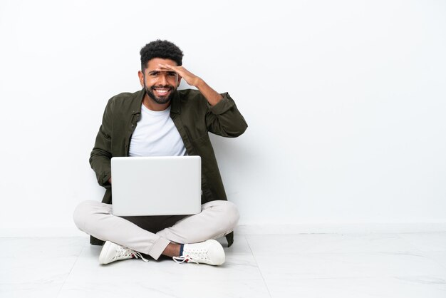 Young brazilian man with a laptop sitting on the floor isolated\
on white looking far away with hand to look something