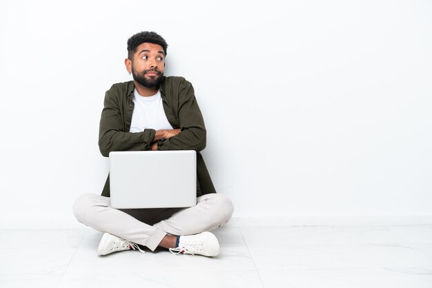 Young Brazilian man with a laptop sitting on the floor isolated on white making doubts gesture while lifting the shoulders