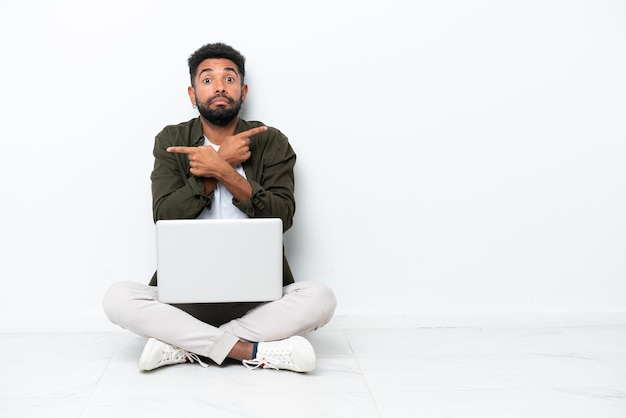 Giovane brasiliano con un laptop seduto sul pavimento isolato su bianco che indica i laterali che hanno dubbi