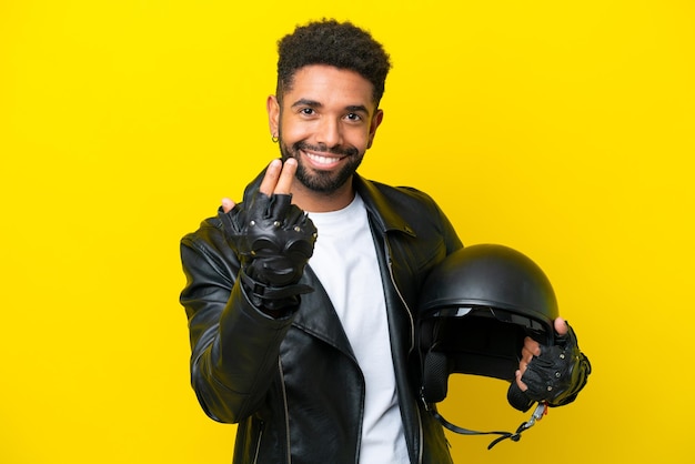 黄色の背景に分離されたオートバイのヘルメットを持った若いブラジル人男性が手で来るように誘うあなたが来たことを幸せに