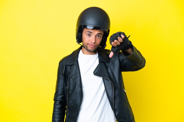 Молодой бразилец в мотоциклетном шлеме на желтом фоне показывает большой палец вниз с негативным выражением лица
