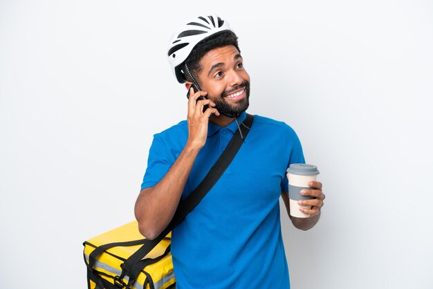 Young Brazilian man with thermal backpack isolated on white background holding coffee to take away and a mobile