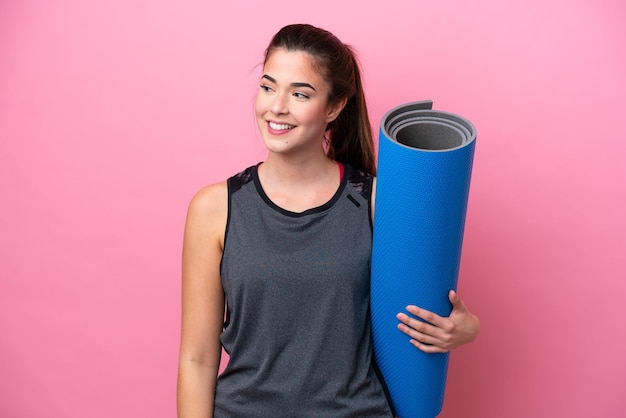 Young Brazilian sport woman going to yoga classes while holding a mat isolated on pink background looking to the side and smiling