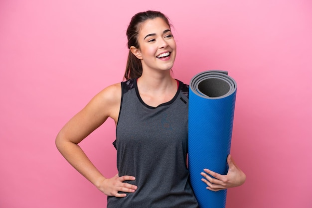 Young Brazilian sport woman going to yoga classes while holding a mat isolated on pink background posing with arms at hip and smiling