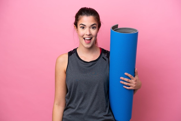 Giovane donna sportiva brasiliana che va a lezioni di yoga mentre tiene un tappetino isolato su sfondo rosa con espressione facciale a sorpresa