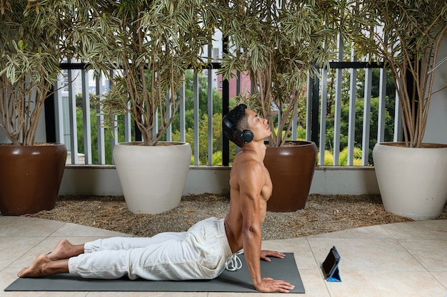 Foto giovane brasiliano che tiene una lezione di yoga online con la cuffia, nella posizione yoga del cane a faccia in su