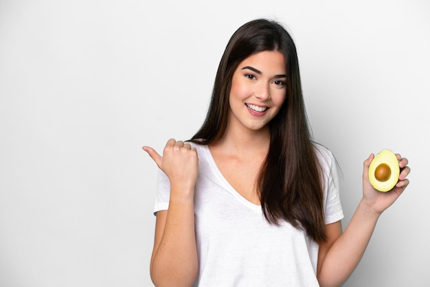 Young Brazilian woman holding an avocado isolated on white background pointing to the side to present a product