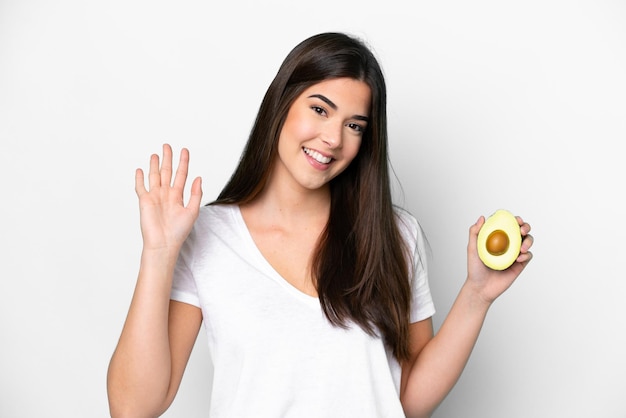 Young Brazilian woman holding an avocado isolated on white background saluting with hand with happy expression