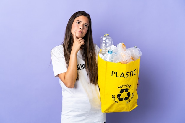 Молодая бразильская женщина, держащая сумку, полную пластиковых бутылок для переработки, изолирована на фиолетовом и сомневается, глядя вверх