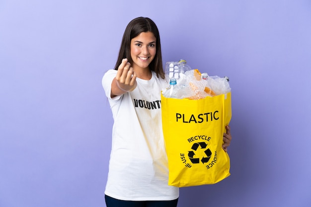 Photo young brazilian woman holding a bag full of plastic bottles to recycle isolated on purple making money gesture