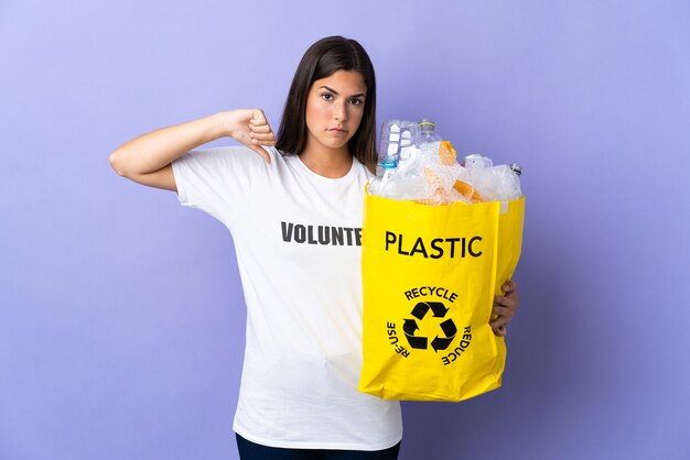 Young brazilian woman holding a bag full of plastic bottles to recycle on purple showing thumb down