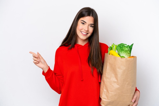 Young Brazilian woman holding a grocery shopping bag isolated on white background pointing finger to the side