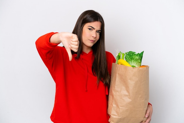 Young Brazilian woman holding a grocery shopping bag isolated on white background showing thumb down with negative expression