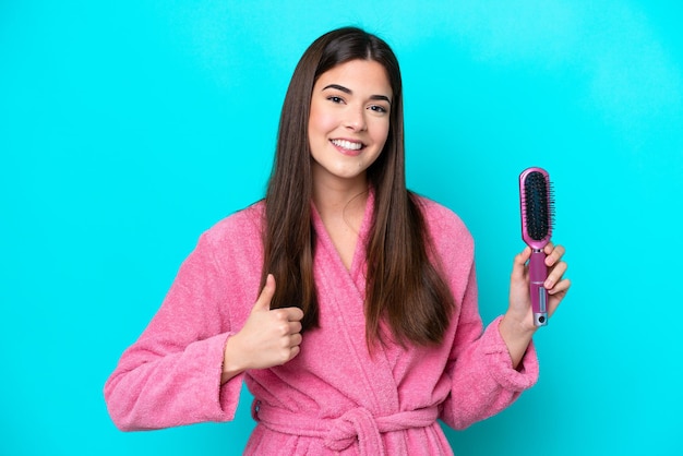 Young Brazilian woman holding hairbrush isolated on blue background with thumbs up because something good has happened
