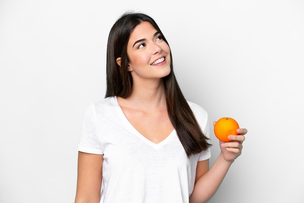 Young Brazilian woman holding an orange isolated on white background looking up while smiling