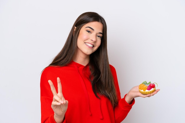 Young Brazilian woman holding a tartlet isolated on white background smiling and showing victory sign